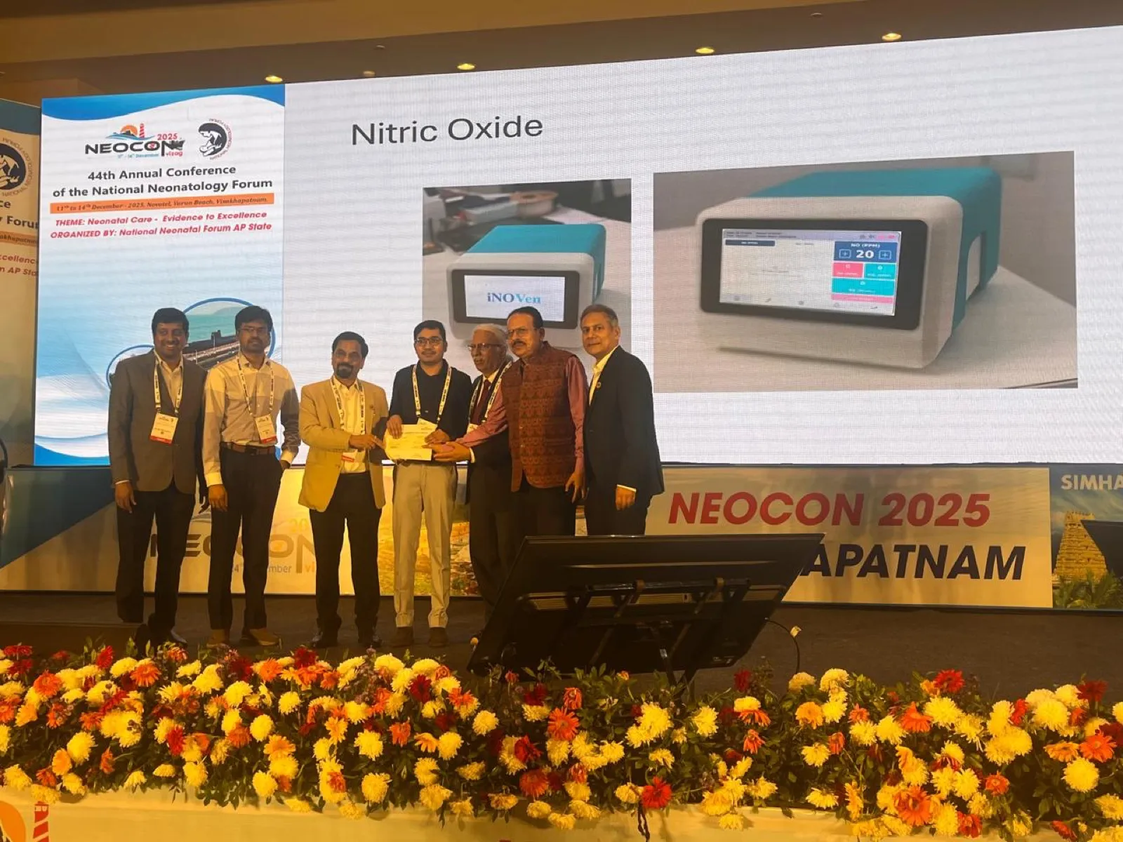The VEHA team receiving recognition for iNO-VEN on stage at NEOCON 2025 in Visakhapatnam, with the device shown on the screen behind them